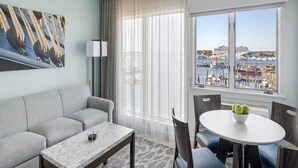 A modern hotel room with a grey sofa, a marble coffee table, a dining table with chairs, a large window with a view of a marina and boats.