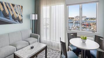 A modern hotel room with a grey sofa, a marble coffee table, a dining table with chairs, a large window with a view of a marina and boats.