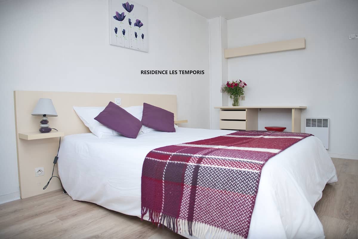 A modern bedroom with a bed, a bedside table, a lamp, and a wall-mounted shelf with a vase of flowers.
