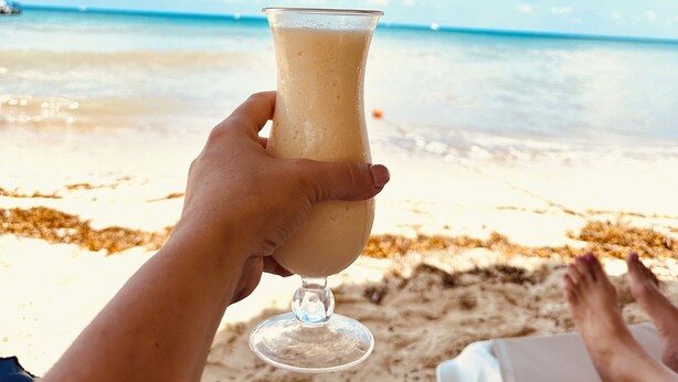 A hand holding a glass of drink with a beach and ocean in the background. - Cozumel-Beautiful 2 bedroom ocean view condo (San Miguel de Cozumel)