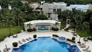 A large outdoor swimming pool with sun loungers and a pergola, surrounded by lush greenery and a view of a residential area. - Cozumel-Beautiful 2 bedroom ocean view condo (San Miguel de Cozumel)