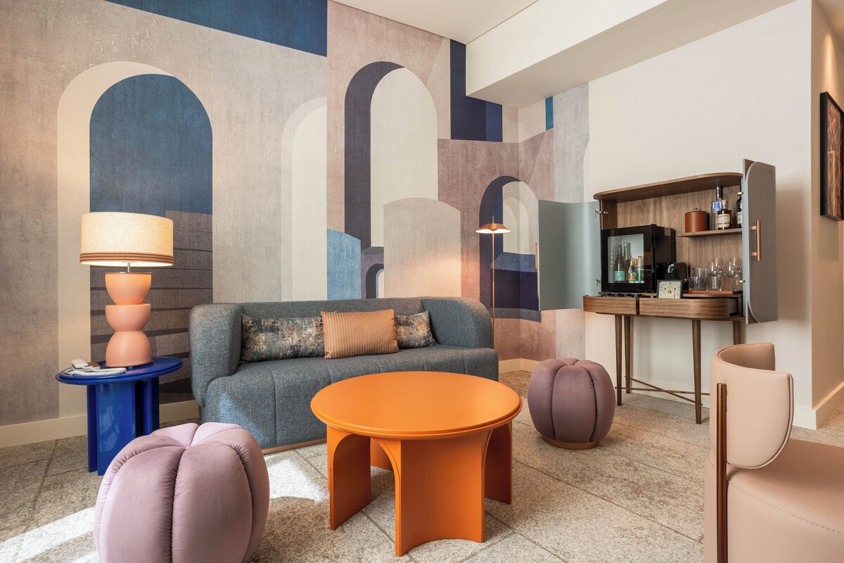 A modern living room with a sofa, round orange table, and a coffee table. There is a small side table with a lamp, a purple ottoman, and a chair. The room has a large abstract wall art and a wooden cabinet with glassware.