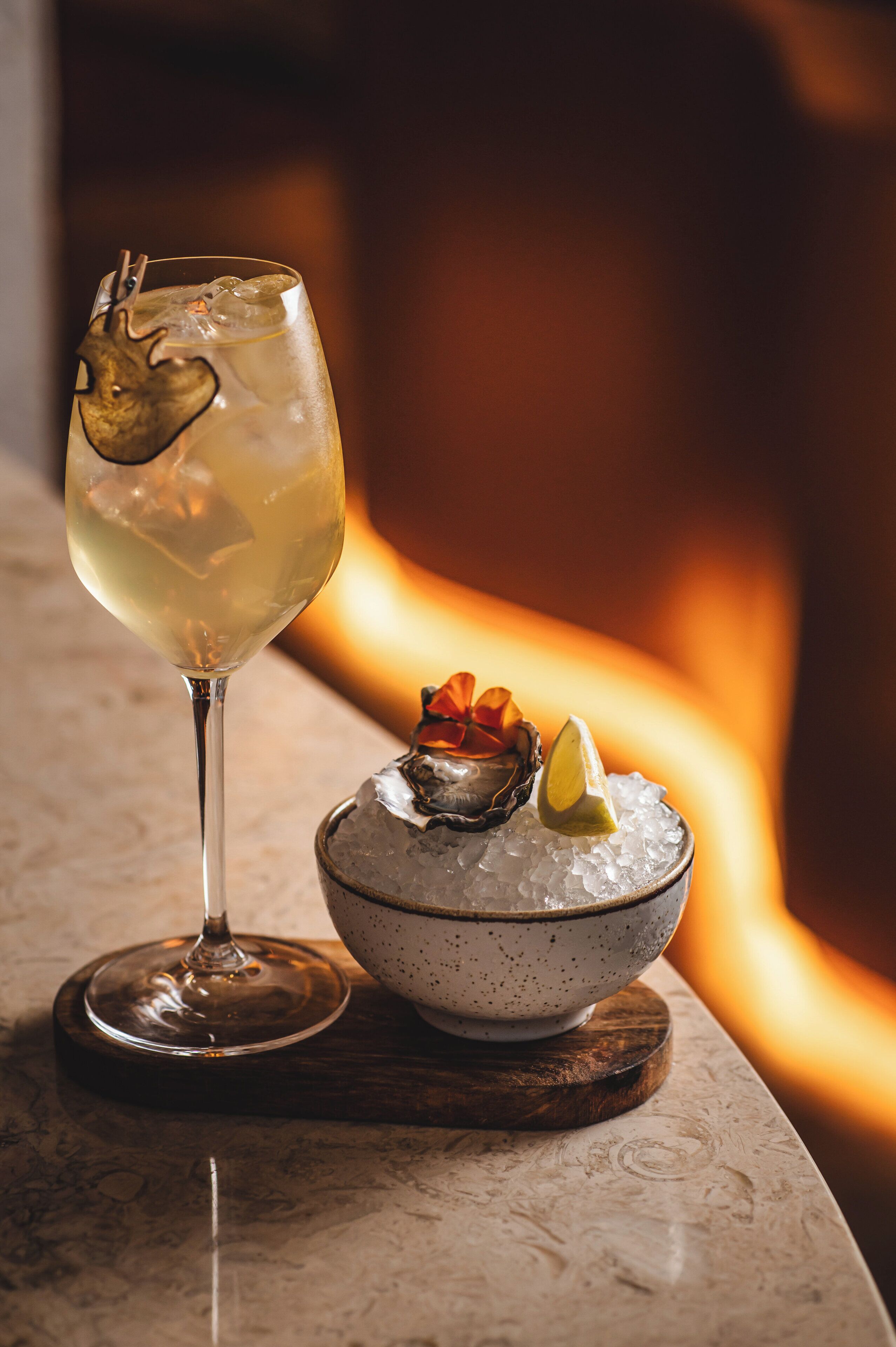 A glass of white wine with a heart-shaped garnish, a bowl of ice with an oyster and a lemon slice, and a wooden coaster.
