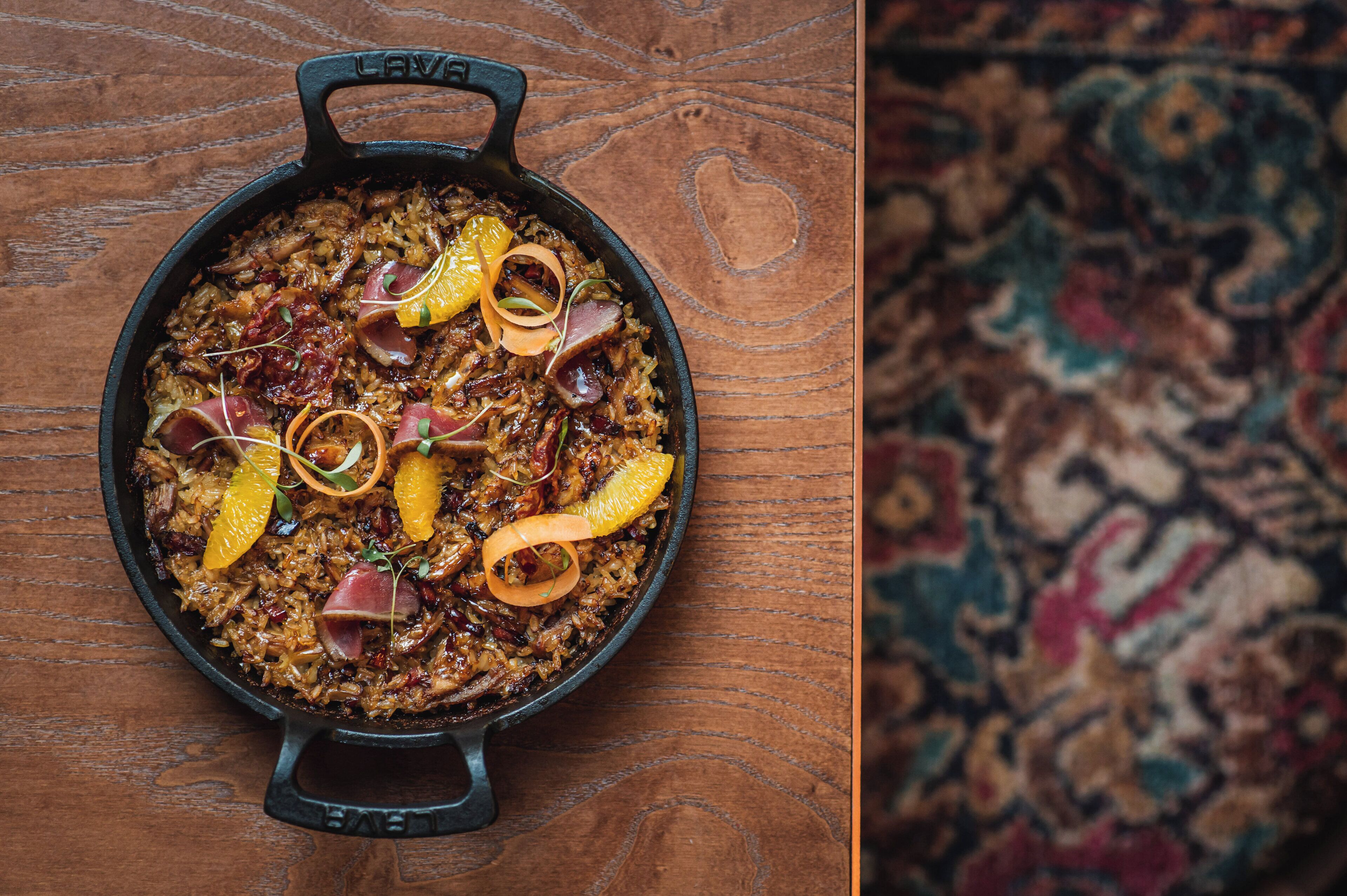 Una sartén de hierro fundido llena de un plato adornado con rodajas de naranja y hierbas, sobre una superficie de madera.
