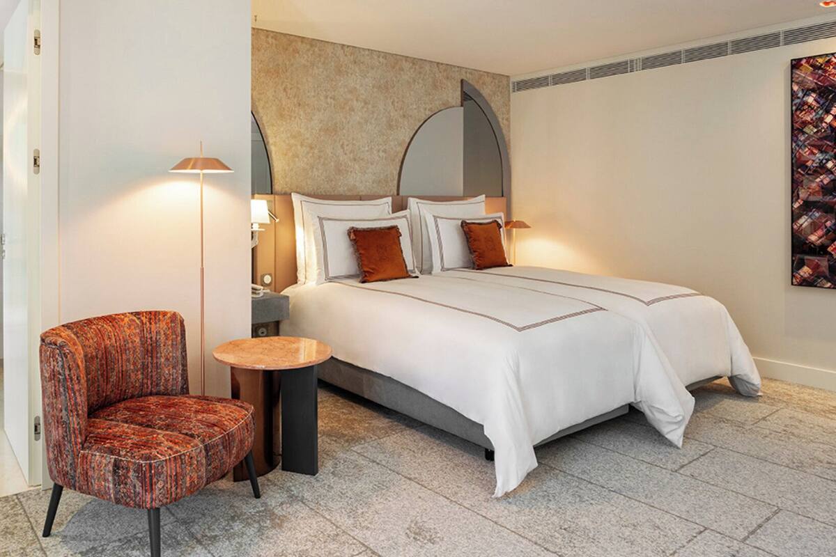 A modern hotel room with a large bed, a round mirror, a small round table, a chair, and a bathroom with a vanity and towel rack.