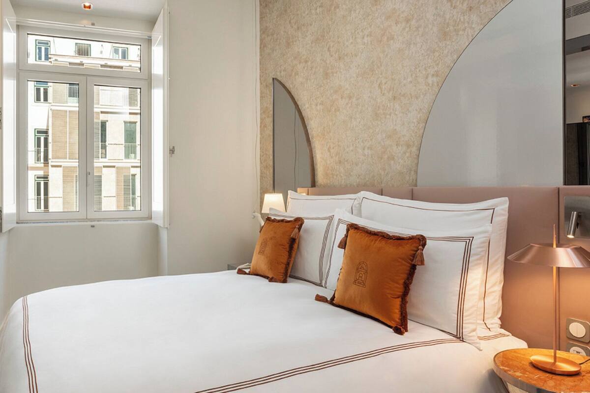 A hotel room with a large bed, a window with a view of buildings, a bedside table with a lamp, and a chair.