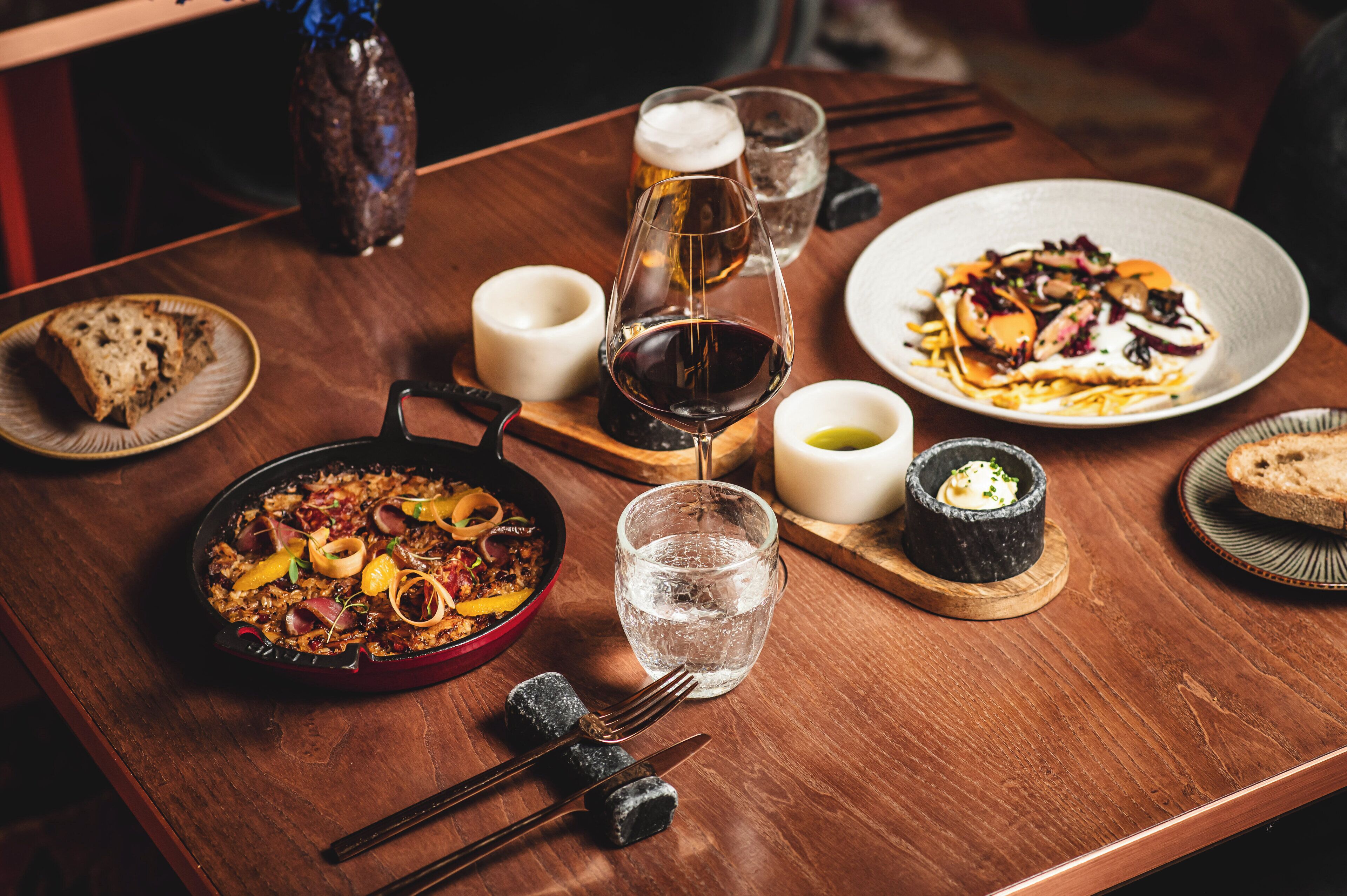 A wooden table with a variety of dishes, including a pan of food, a plate of vegetables, and a bowl of soup. There are also glasses of water and a bottle of beer.