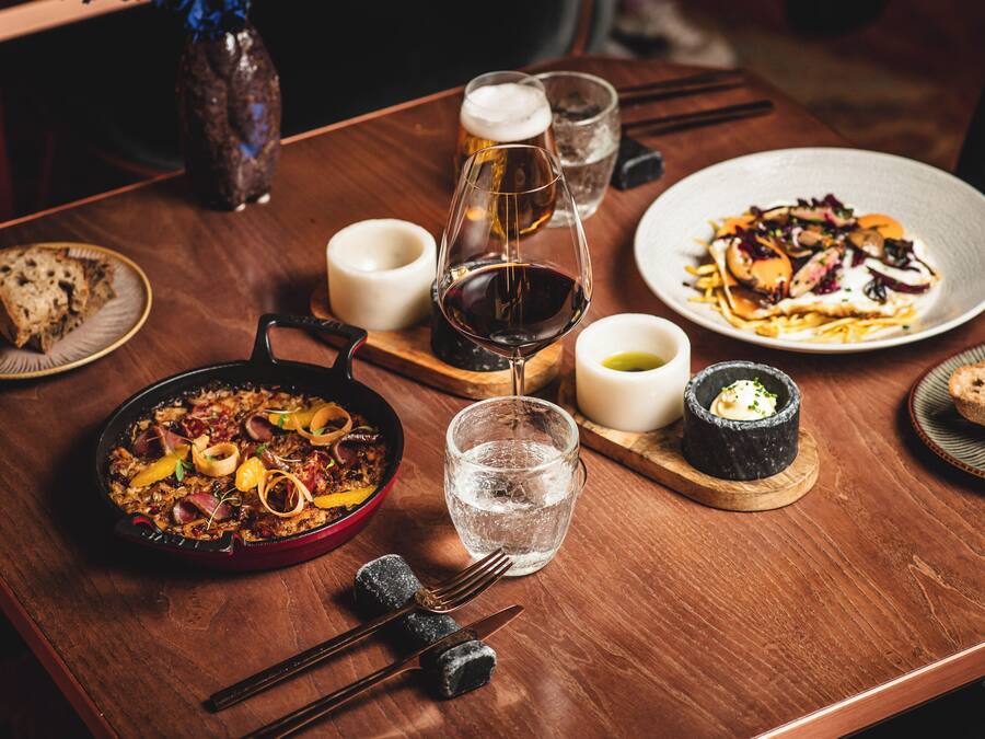 Una mesa de madera con una variedad de platos, incluyendo una paellera con comida, un plato de verduras y un tazón de sopa. También hay vasos de agua y una botella de cerveza.