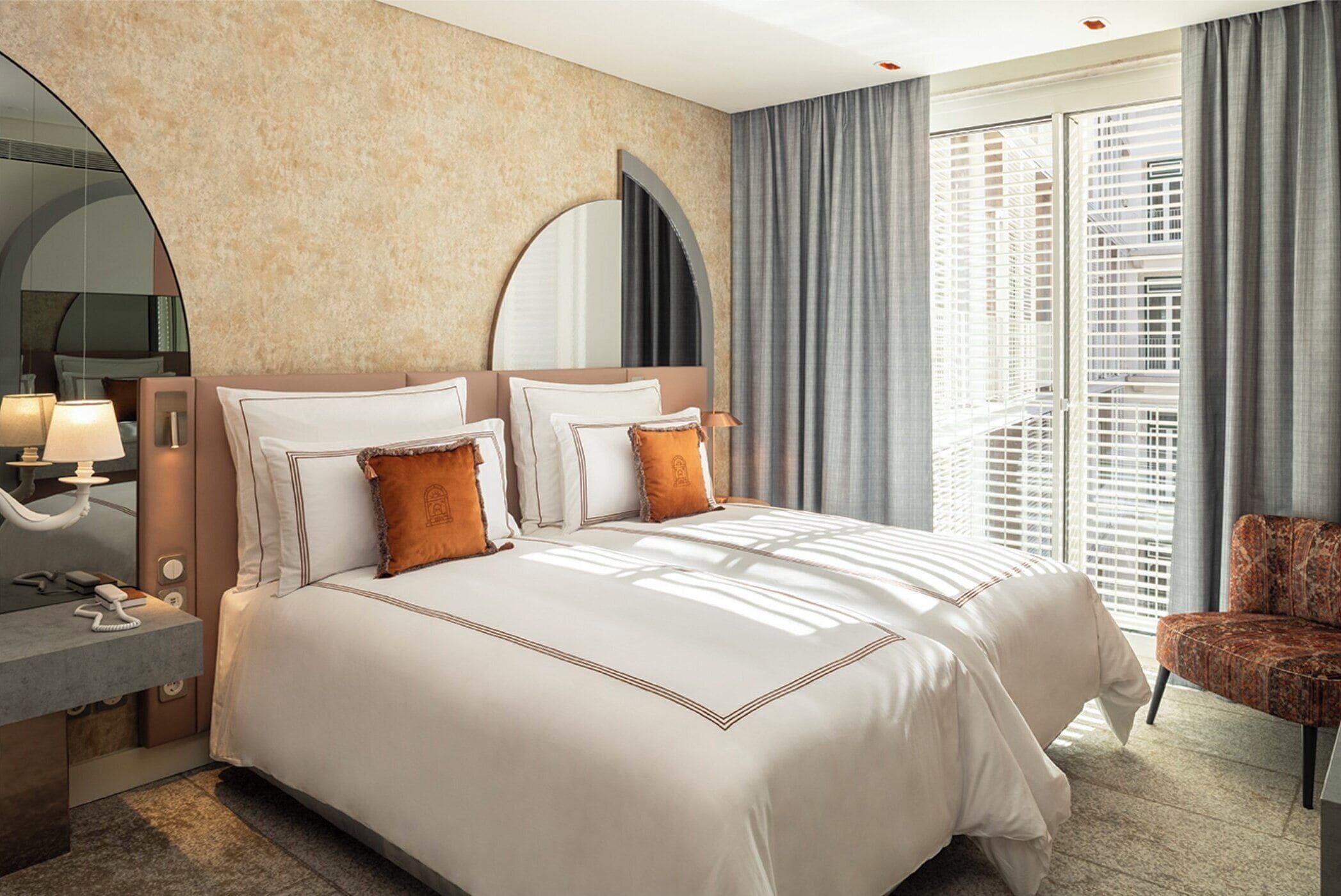 Quarto de hotel com uma cama grande, uma mesa de cabeceira com abajur, uma cadeira e uma janela ampla com persianas.
