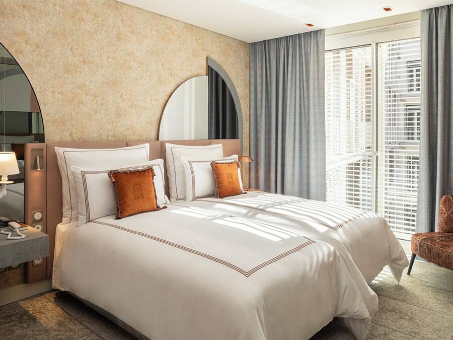 Quarto de hotel com uma cama grande, uma mesa de cabeceira com abajur, uma cadeira e uma janela ampla com persianas.