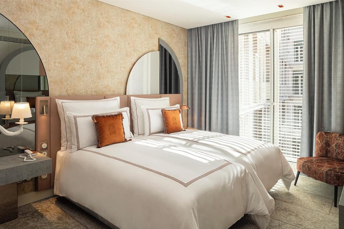 A hotel room with a large bed, a nightstand with a lamp, a chair, and a large window with blinds.