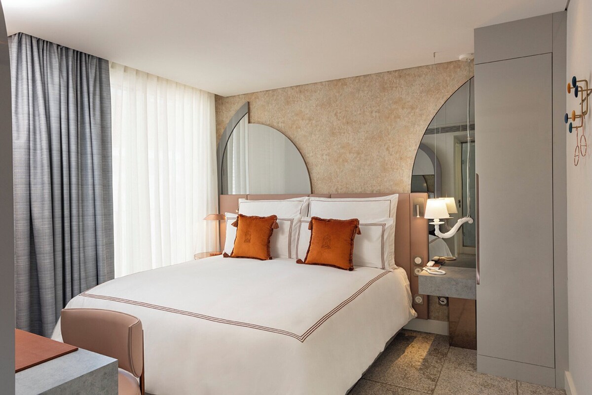 A hotel room with a large bed, two pillows, a nightstand, a lamp, a mirror, and a wardrobe.