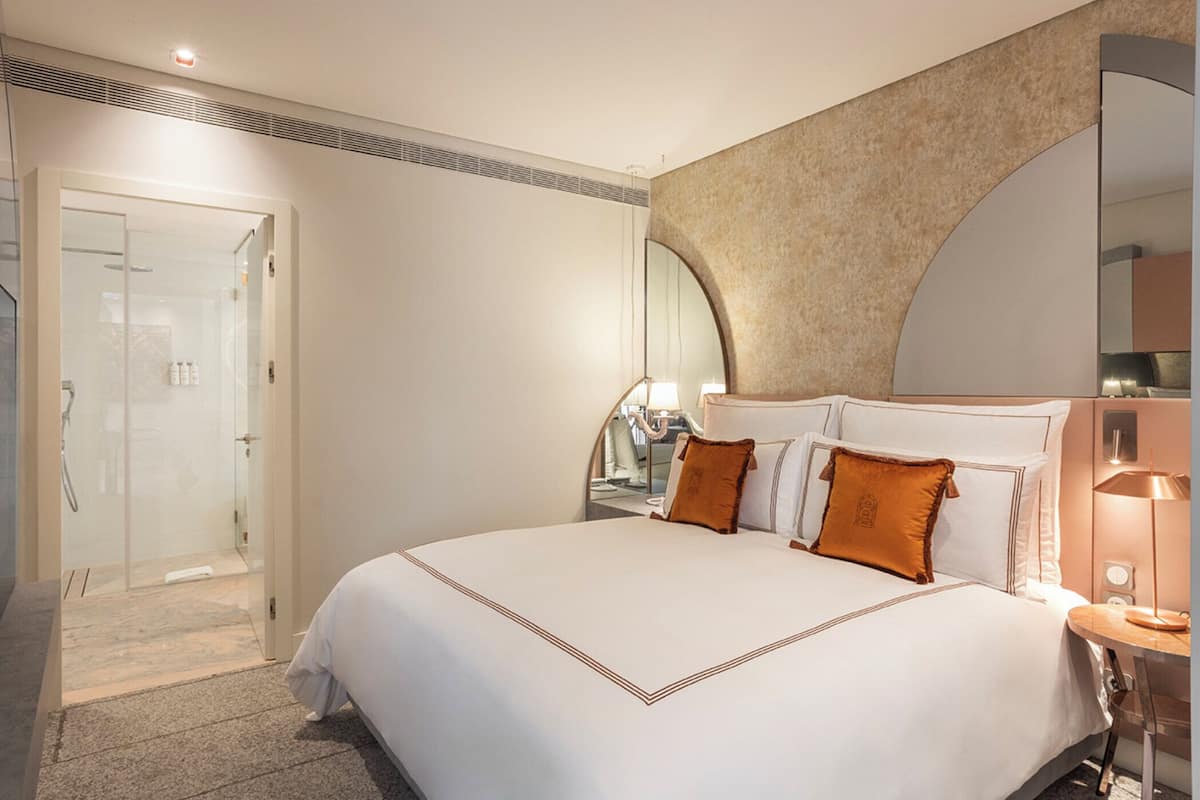 A modern hotel room with a large bed, a glass shower enclosure, and a curved mirror above the bed.