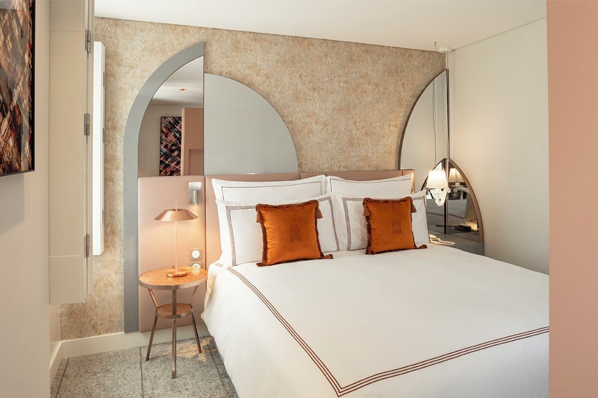 A hotel room with a large bed, a round table, a lamp, and a mirror.