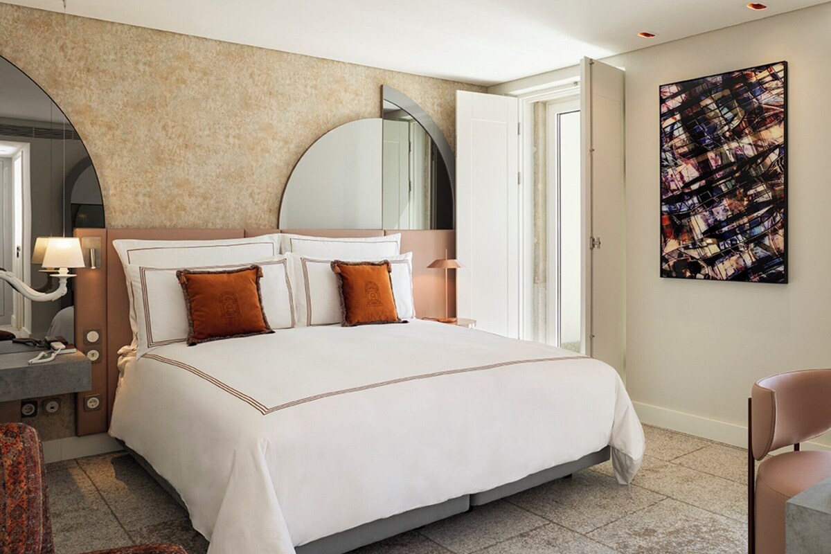 A hotel room with a large bed, a sitting area with a chair, a desk with a lamp, and a large abstract painting on the wall.