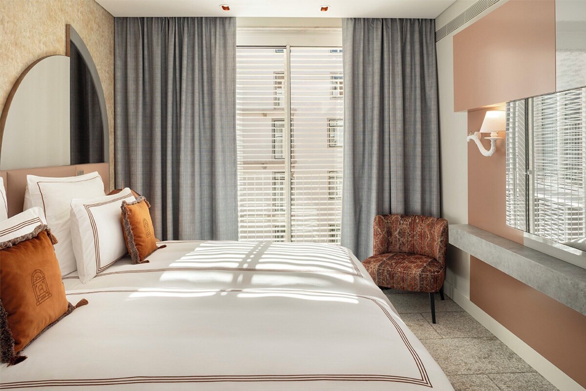 A hotel room with a large bed, a chair, a window with blinds, and a wall-mounted lamp.