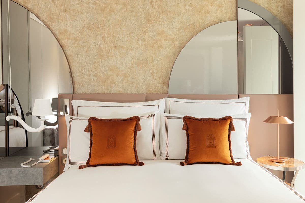 A hotel room with a large bed, two pillows, a bedside table with a lamp, and a mirror above the bed.