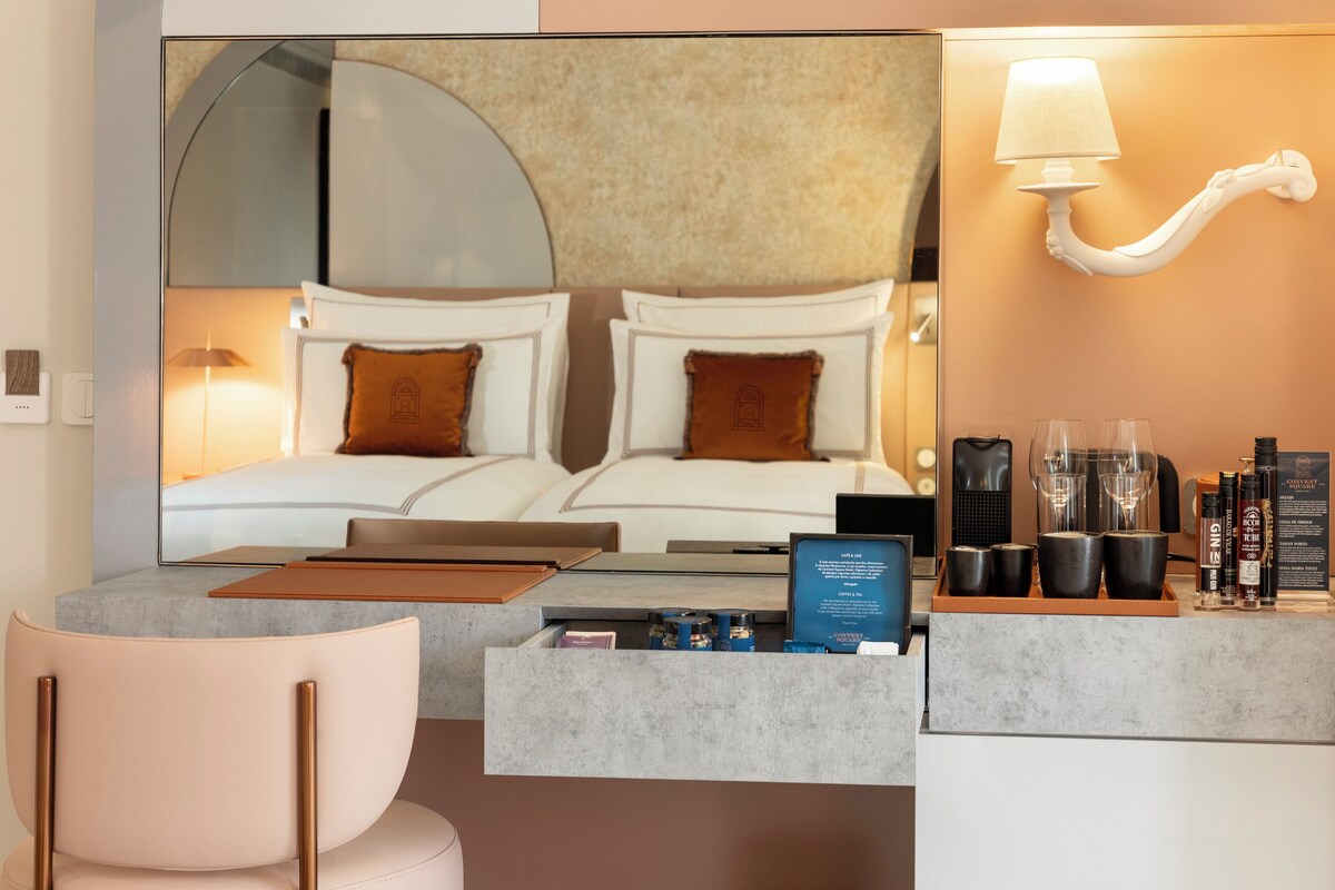 A modern hotel room with a large bed, a desk with a laptop, a coffee machine, and a tray of toiletries.