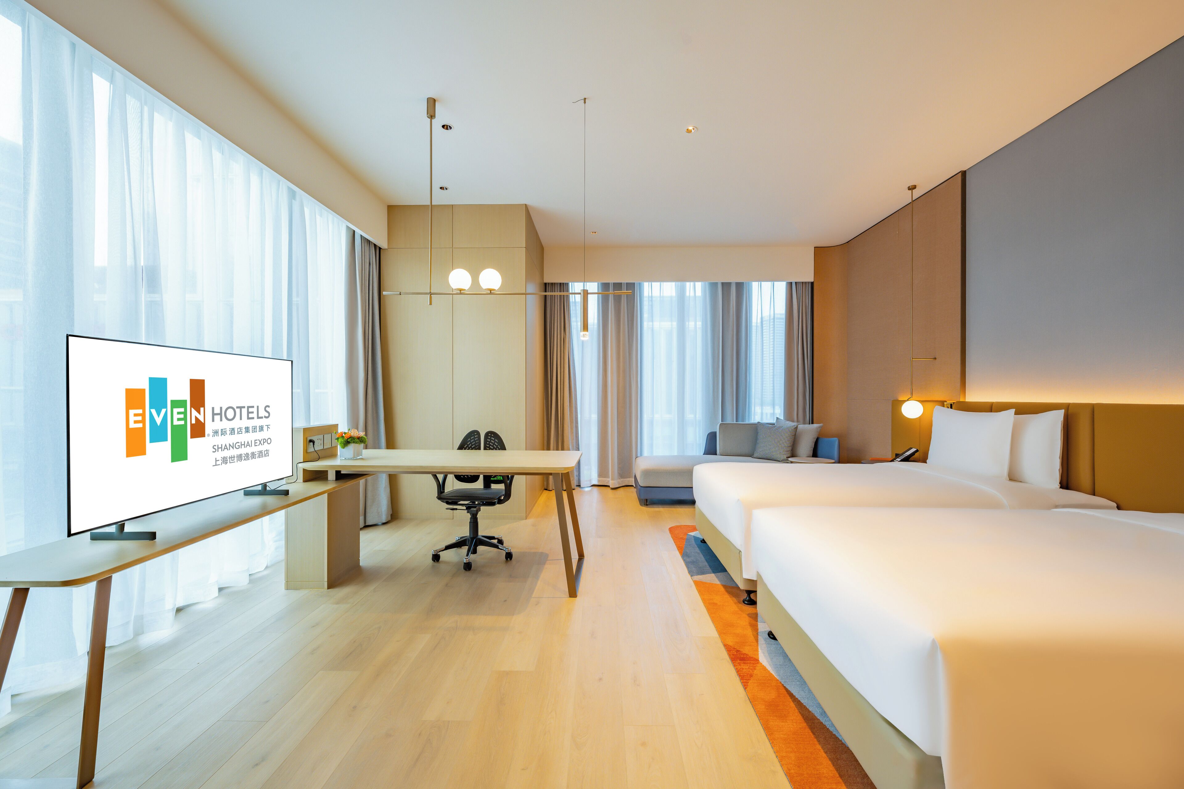 A modern hotel room with a large bed, a desk, a chair, and a television displaying the hotel's name.