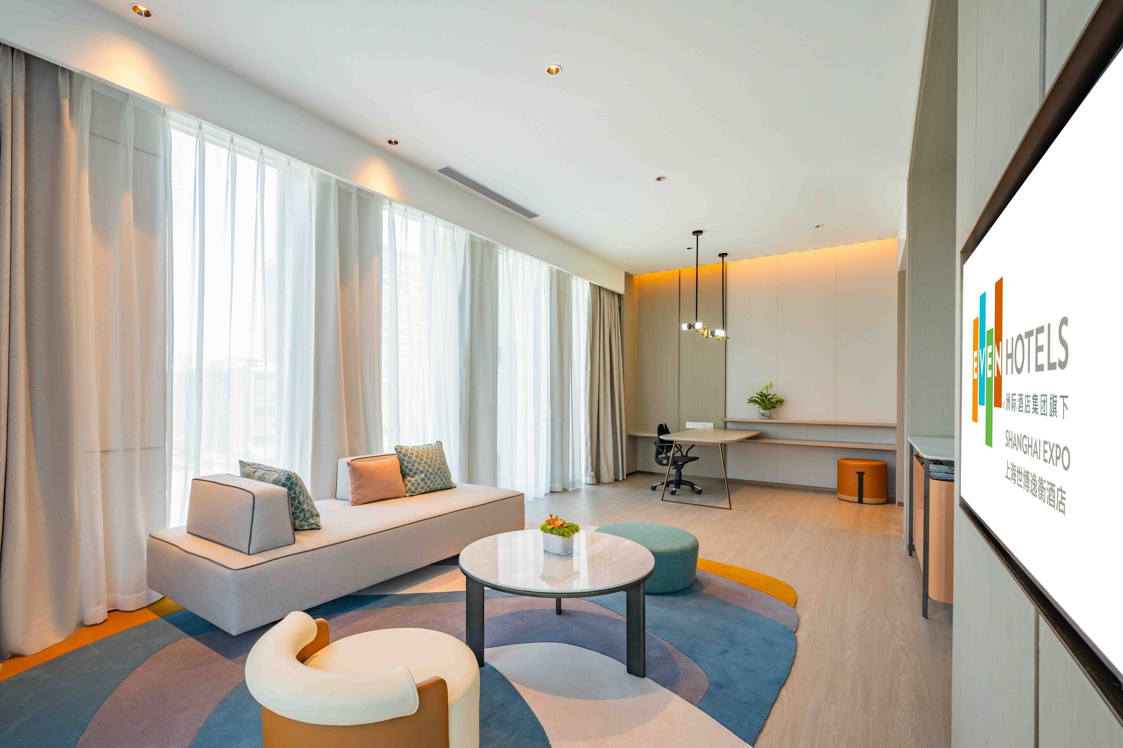 A modern hotel room with a large window, a sofa, a round coffee table, and a small desk.