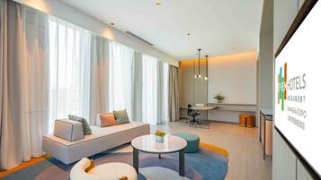 A modern hotel room with a large window, a sofa, a round coffee table, and a small desk.