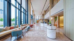 Een ruime lobby met een lange receptiebalie, een zithoek met banken en stoelen, en grote ramen met uitzicht op buiten.