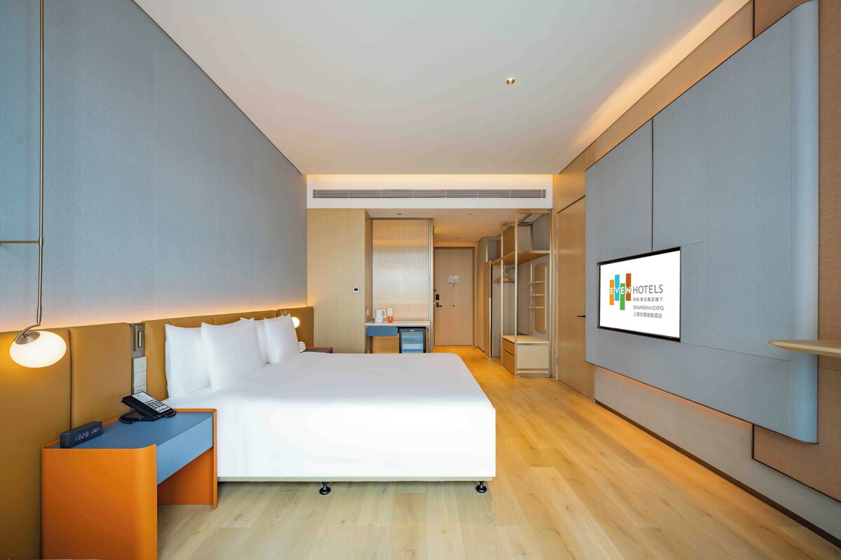 A modern hotel room with a large bed, bedside table with a phone, a desk with a computer, and a TV displaying the hotel name.