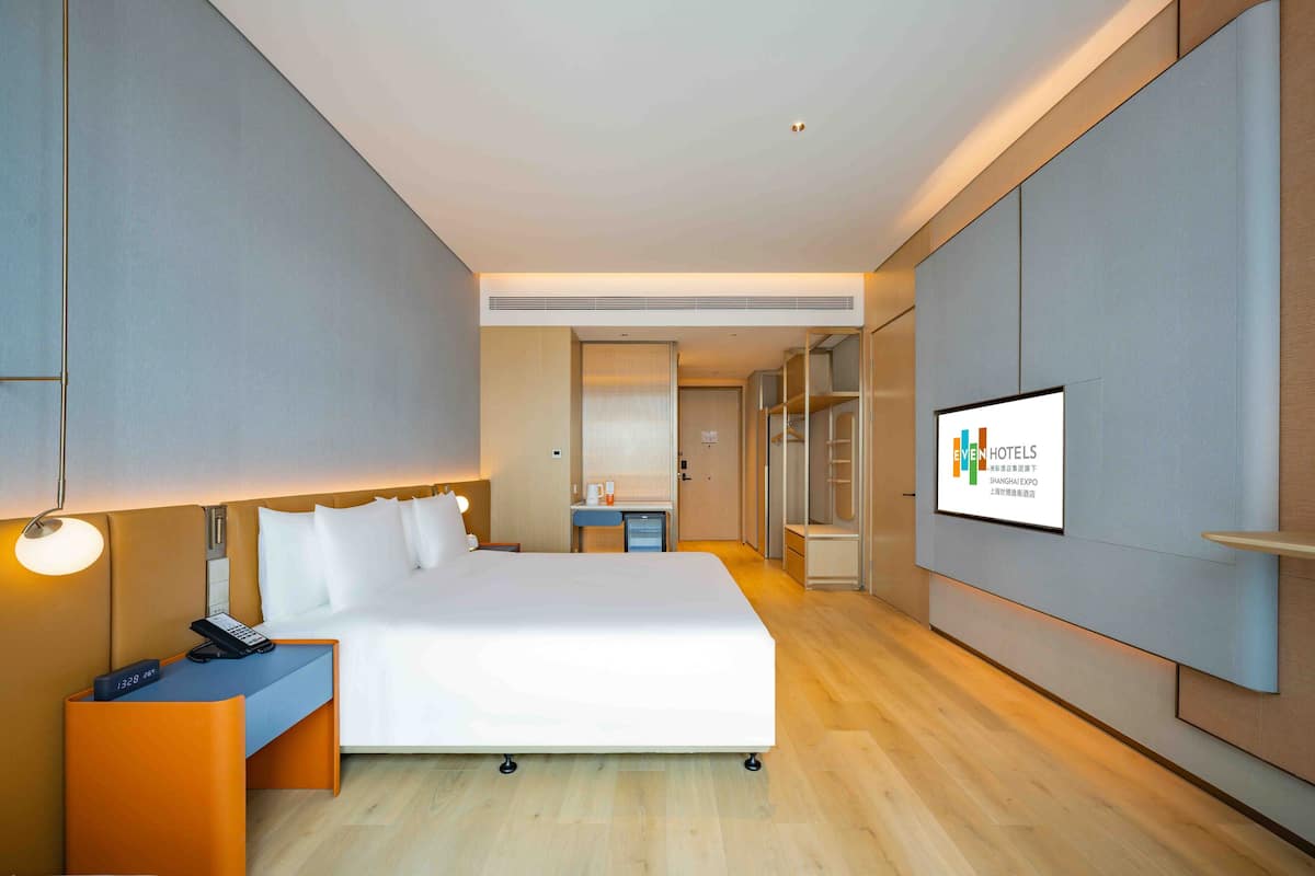 A modern hotel room with a large bed, bedside table with a phone, a desk with a computer, and a TV displaying the hotel name.