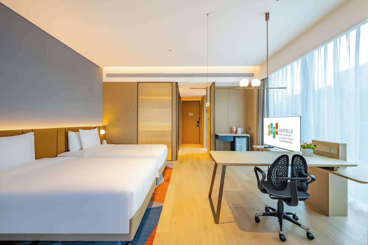 A hotel room with two beds, a desk, a chair, and a large window.