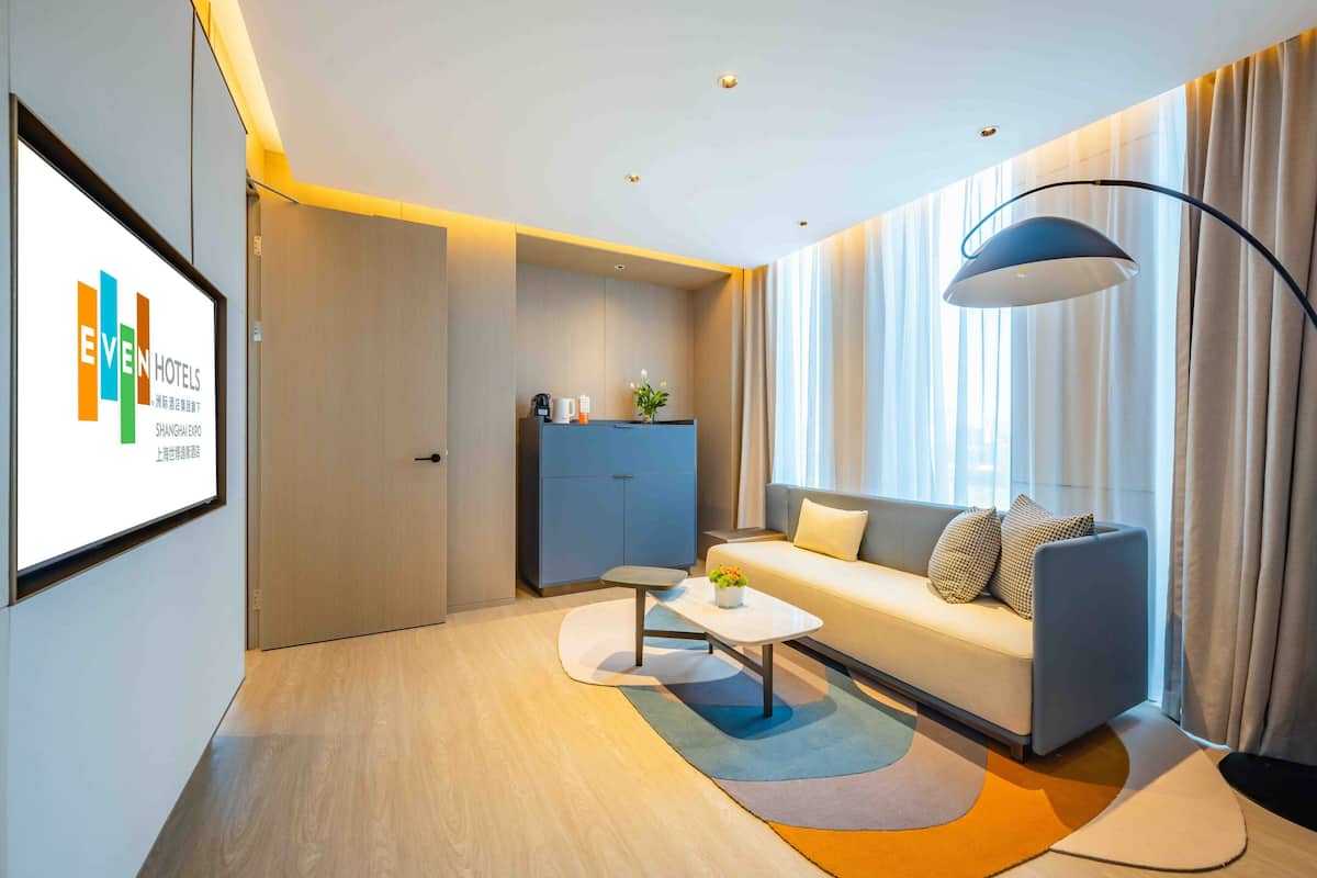 A modern hotel room with a sofa, a coffee table, a lamp, and a view of the city.