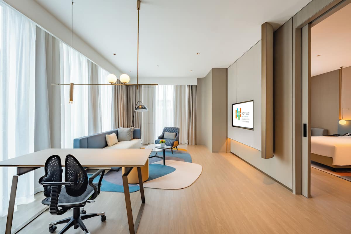 A modern hotel room with a large bed, a desk with two chairs, a sofa, and a TV screen displaying a hotel sign.