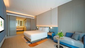 Een moderne hotelkamer met een groot bed, een bank, een klein tafeltje met een plant, en een badkamer zichtbaar door een schuifdeur.