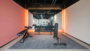 Een fitnessruimte met trainingsapparatuur, waaronder gewichten en banken, en een vloerbedekking.