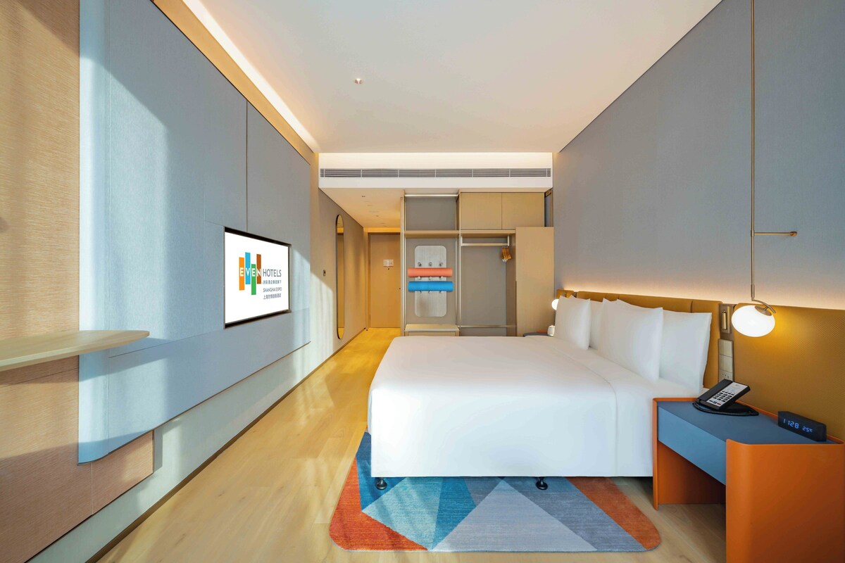 A modern hotel room with a large bed, bedside table with a phone, and a wall-mounted TV.