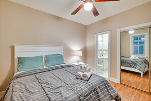 A bedroom with a bed, a ceiling fan, a nightstand with a lamp, a bathroom mirror, and a view of the bathroom. - Golf, Swim & Unwind at Bright Myrtle Beach Condo! (Myrtle Beach)