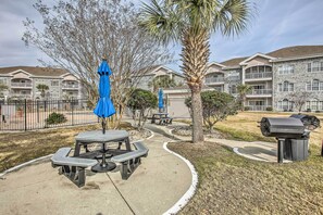 A courtyard with picnic tables and blue umbrellas, surrounded by a fence and a building with balconies. - Golf, Swim & Unwind at Bright Myrtle Beach Condo! (Myrtle Beach)