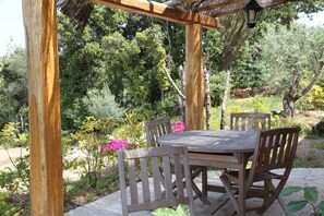 An outdoor wooden pergola with a table and chairs, surrounded by lush greenery and flowering plants.