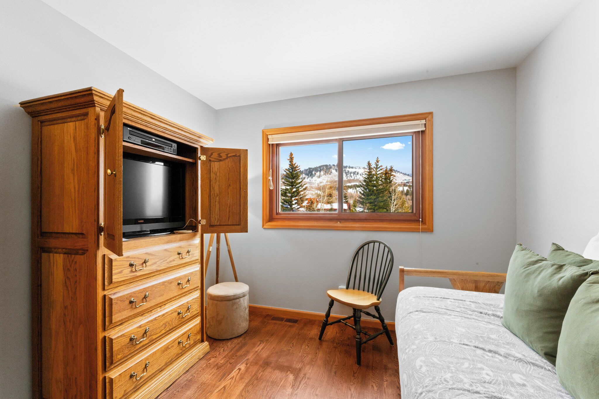 Een slaapkamer met een houten dressoir, een televisie, een raam met uitzicht op de bergen, een stoel en een bed met groene kussens.