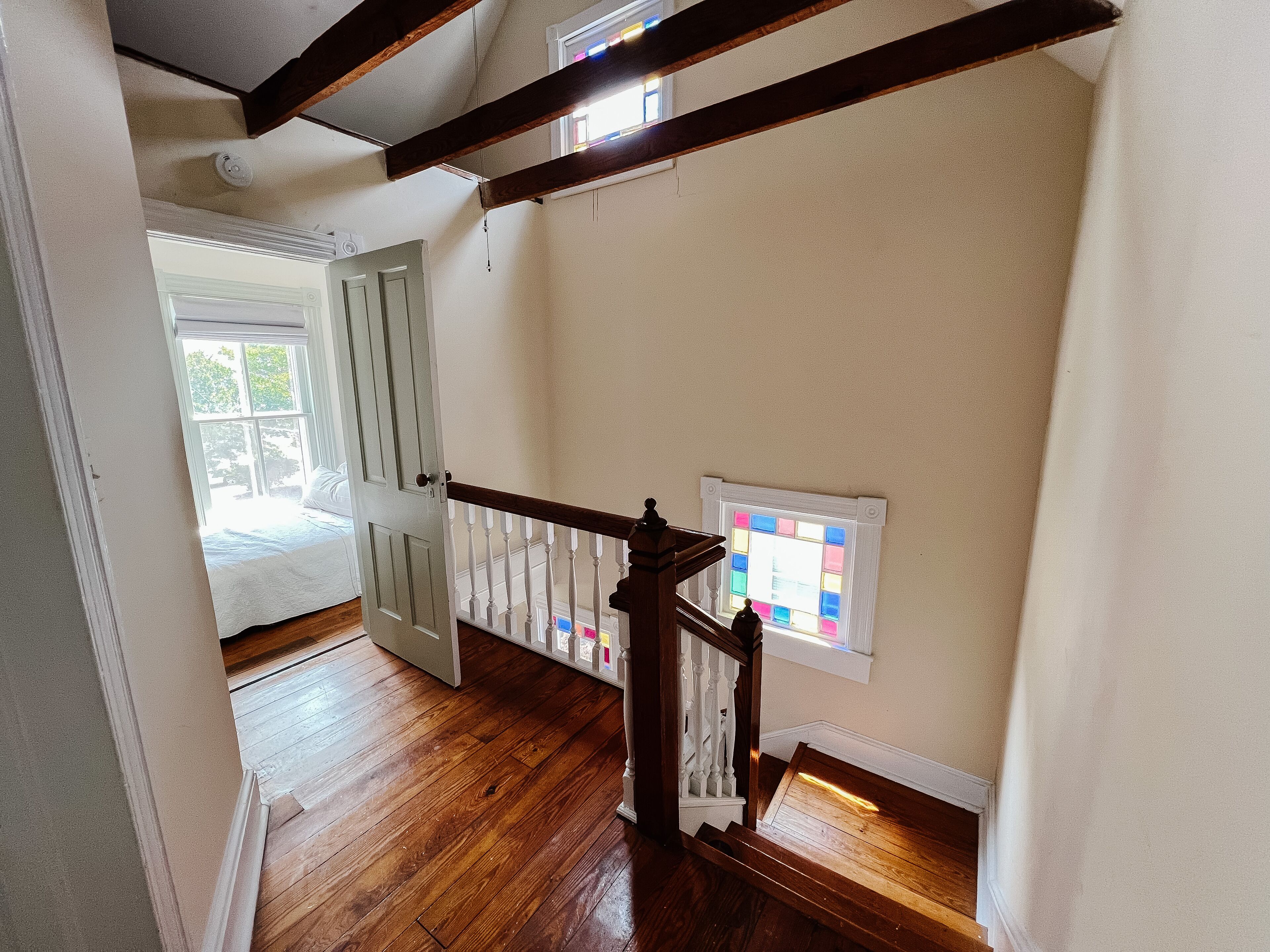 A wooden staircase with a white railing, a door leading to a room with a bed, and a window with colorful panes.