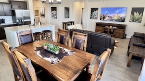 Een moderne keuken met een centraal eiland, een eethoek met een houten tafel en stoelen, een woonkamer met een grote televisie en zithoek, en een plafondventilator.