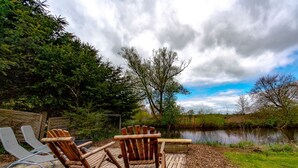 A wooden deck with two Adirondack chairs facing a calm river, surrounded by lush greenery and a cloudy sky.