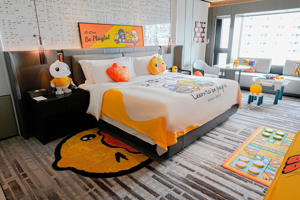 A hotel room with a bed, a nightstand, a window, and a view of the city.