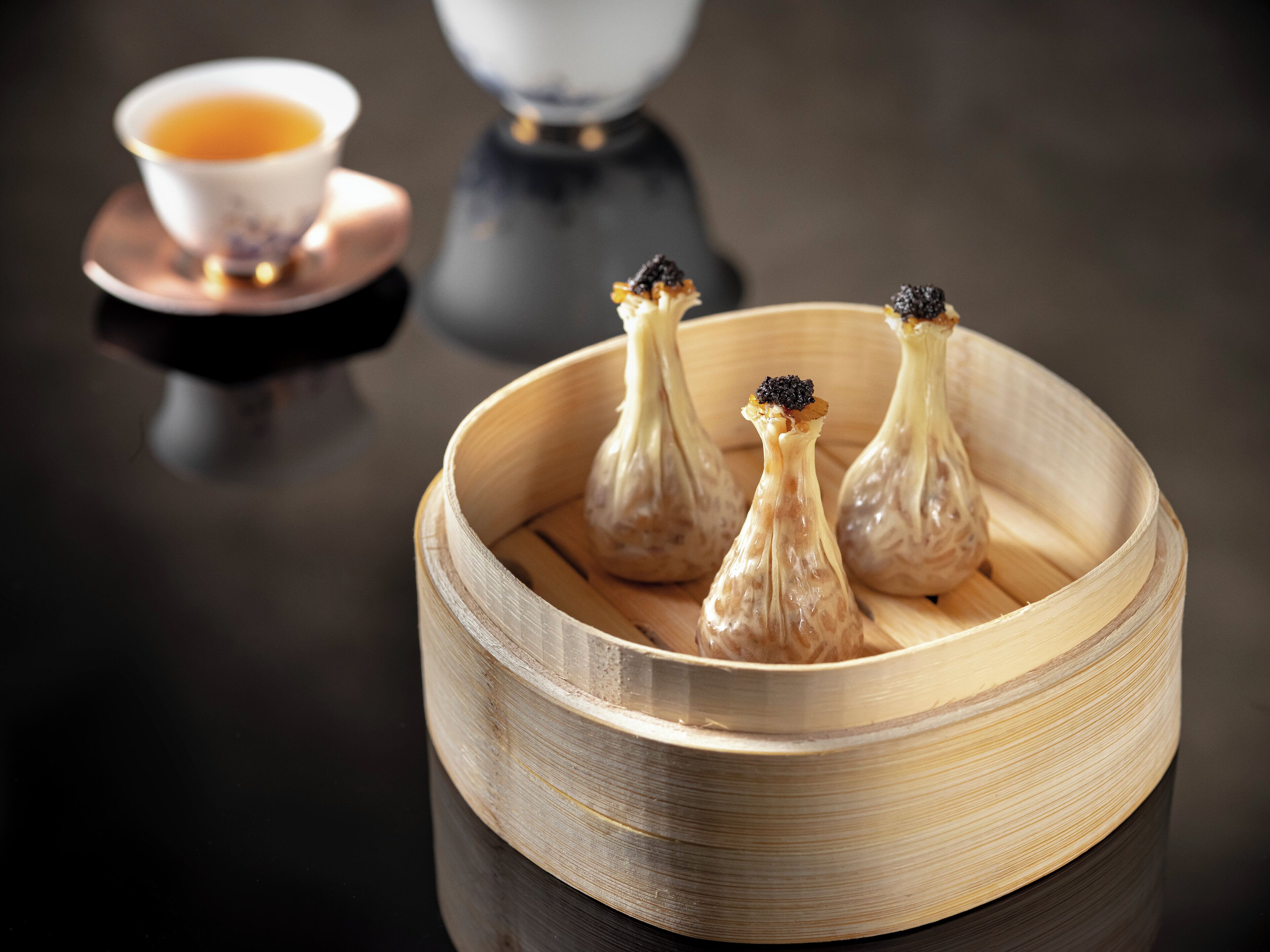 A bamboo steamer with three dumplings, a teapot, and a cup of tea on a reflective surface.