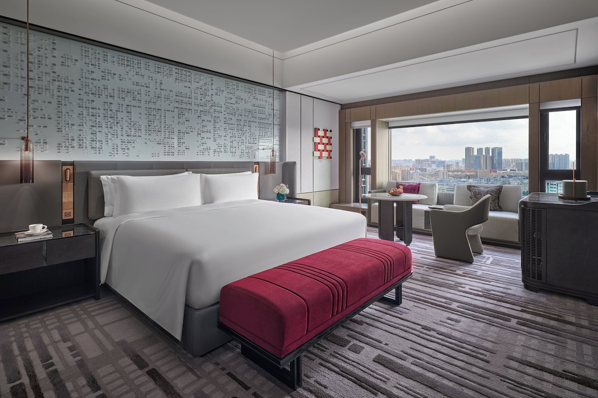 A modern hotel room with a large bed, a red bench, a seating area with a view of the city, and a large window.