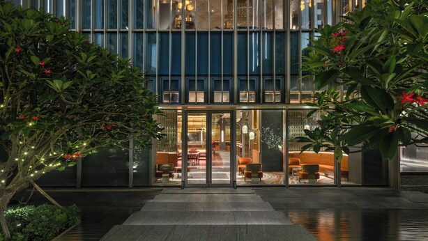 Un edificio moderno con fachada de vidrio, una entrada central con puertas de cristal y zonas de estar interiores visibles a través del vidrio.