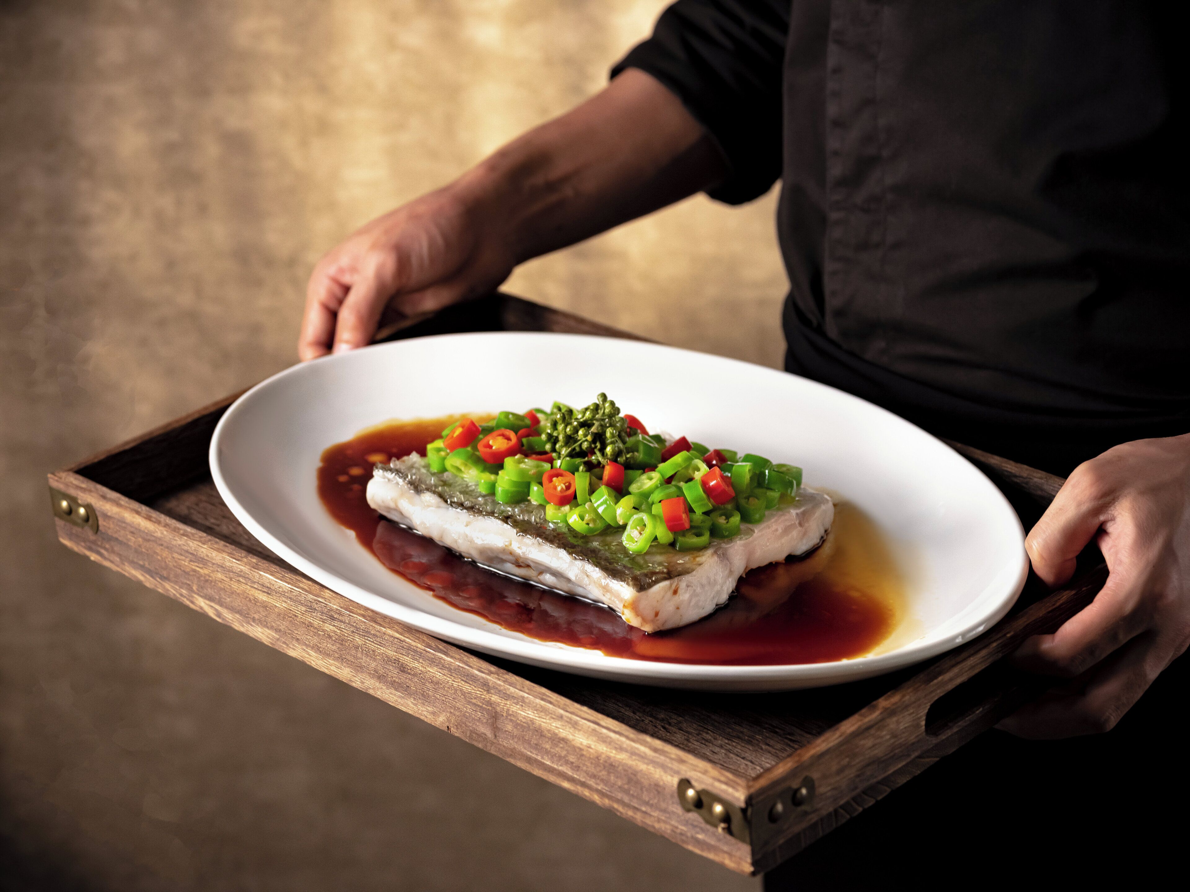 Une personne tient un plateau en bois sur lequel se trouve une assiette blanche contenant un plat de poisson, agrémenté de poivrons verts et rouges, et nappé d’une sauce foncée.