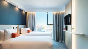 Quarto de hotel com duas camas, uma televisão na parede e vista para a paisagem urbana.