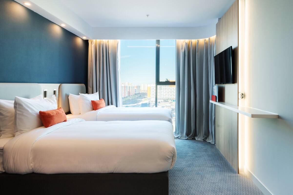 A hotel room with two beds, a TV on the wall, and a view of the cityscape.