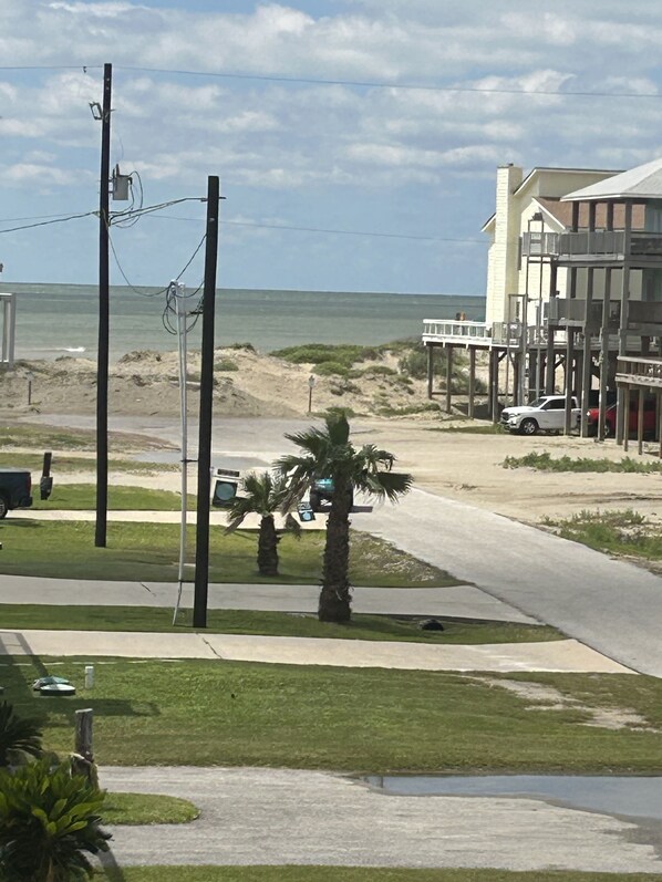 A coastal road with a beach view, a white car parked on the side, and a building with a balcony overlooking the beach.