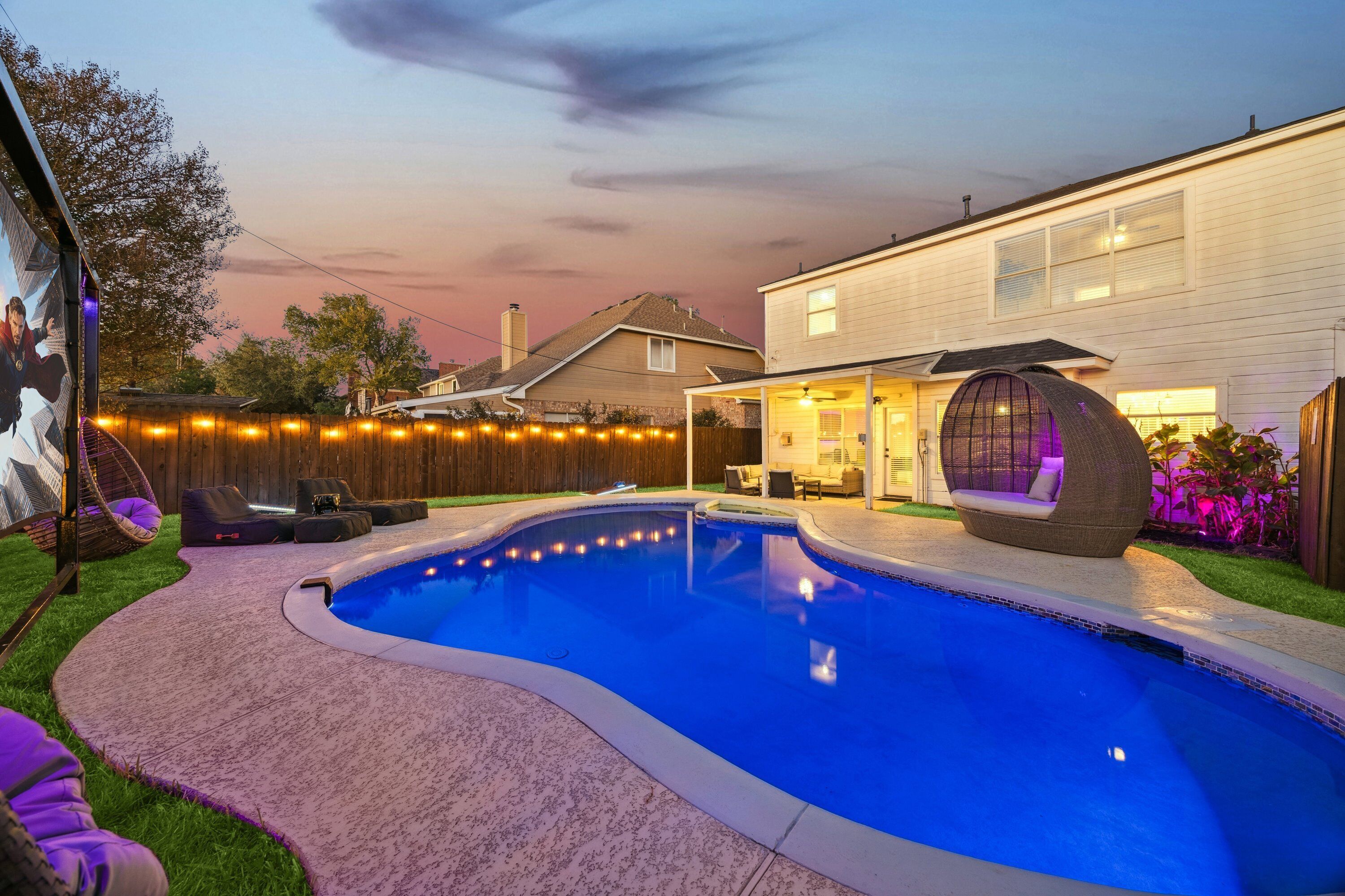 A backyard with a swimming pool, a covered seating area, and a house with a porch.